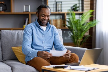 Evdeki kanepede oturan neşeli adam, bir belge okuyor ve yakınlarda bir dizüstü bilgisayarla gülümsüyor..