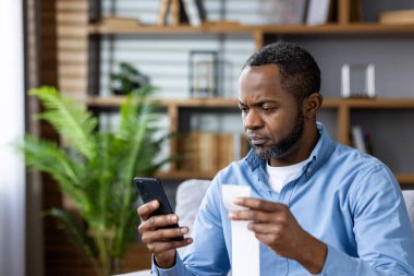 Ciddi bir adam evde otururken telefonundaki uygulamayı kullanarak faturasını ödüyor. Resim dijital ödeme ve mali sorumluluğu betimliyor.
