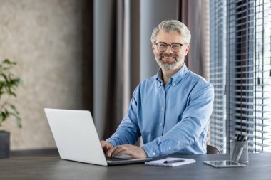 Gülümseyen iş adamı, modern bir ofisin içinde büyük pencereleri ve doğal ışığı olan bir dizüstü bilgisayarda çalışıyor..