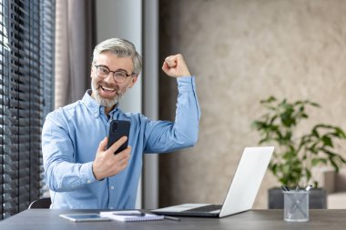 Yaşlı işadamı başarıyı akıllı telefonuyla kutluyor. Gülümsüyor, heyecan gösteriyor ve modern teknolojiyi kullanıyor..