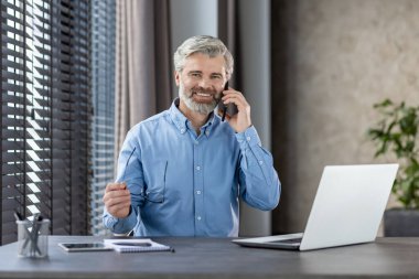 Kendine güveni tam bir iş adamı. Masasında bilgisayarla konuşurken gülümsüyor. Parlak, modern bir ofis alanında..