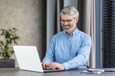 Modern ofis ortamında dizüstü bilgisayar kullanırken gülümseyen gözlüklü ve sakallı yaşlı bir adam.