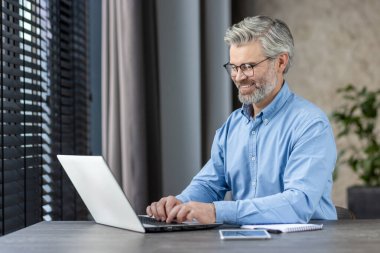 Modern ofis ortamında dizüstü bilgisayarda çalışan gülümseyen olgun iş adamı. Uzaktan çalışma, teknoloji ve meslek hayatı kavramı.