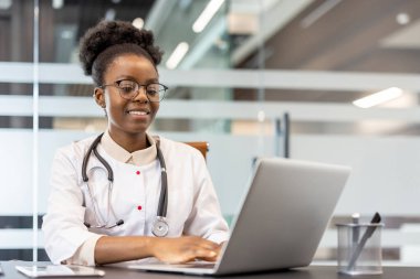 Modern ofis ortamında dizüstü bilgisayar kullanarak gülümseyen bir sağlık uzmanı. Doktor beyaz önlük ve steteskop giyip işine odaklanmış..