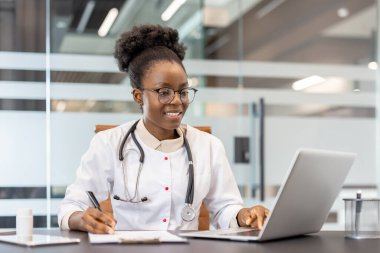Modern bir ofiste dizüstü bilgisayar üzerinde çalışan, notlar alan, steteskop takan, hasta bakımı ve tıbbi araştırmalara odaklanan bir doktor..