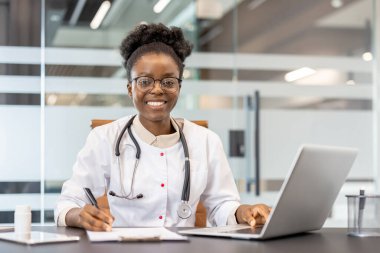 Masada dizüstü bilgisayarla çalışan gülümseyen bir doktor, modern bir muayenehanede notlar yazıyor. Çağdaş bir klinikte profesyonel, sağlık ve teknoloji.