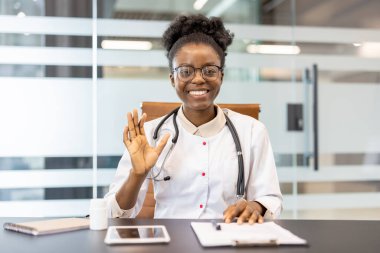 Gülümseyen doktor el sallıyor ve modern ofis ortamında çevrimiçi video görüşmeleri yapıyor. Boynundaki steteskop bir tıp uzmanını işaret ediyor..