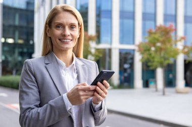 Ofis binasının önünde takım elbiseli, elinde telefonla kameraya güvenle bakan gülümseyen ve başarılı sarışın bir iş kadınının yakın plan portresi..