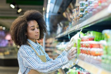İş önlüğü ve tek kullanımlık eldivenli genç Afrikalı Amerikalı kadın süpermarkette çalışıyor, rafların arasında duruyor ve ürünler ve ürünler seriyor..