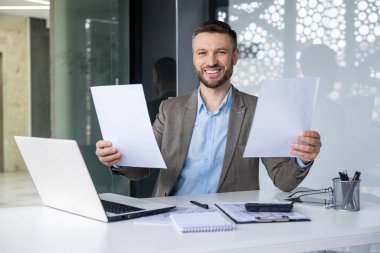 Kendine güvenen güler yüzlü işadamı modern ofis ortamında masasında dokümanları sergiliyor. Laptop ve ofis malzemeleriyle profesyonel çalışma alanı.