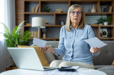 Yaşlı kadın evde bilgisayarını ve faturalarını tutarken stresli ve endişeli görünüyor. Mali zorluklar ve yönetim masrafları kavramı.