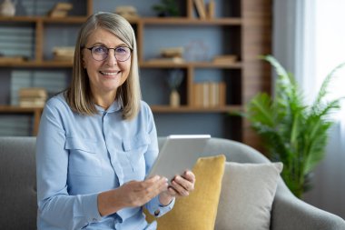 Evde dijital tablet kullanan, modern oturma odasındaki kanepede dinlenen gözlüklü mutlu bir son sınıf kadını. Teknoloji, bağlantı ve rahatlık..