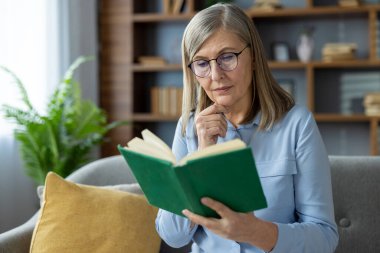 Kanepede oturan gözlüklü yaşlı kadın, evde kitap okumakla çok meşgul..