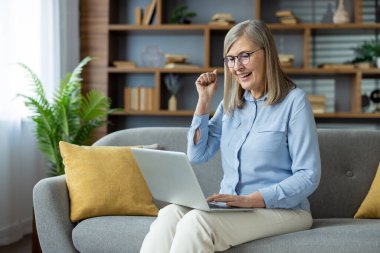 Evde dizüstü bilgisayar kullanırken koltukta oturmuş başarıyı kutlayan son sınıf kadını. Başarılı bir sonuç için olumlu duygular, mutluluk ve heyecan..