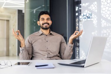 Başarılı İspanyol adam ofiste dinleniyor, iş adamı işyerinde Lotus pozisyonunda meditasyon yapıyor, ara sırasında dizüstü bilgisayarla yoga yapıyor.