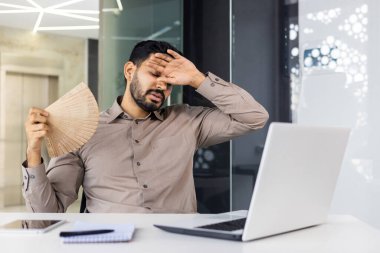 Kahverengi gömlekli bir işadamı, bir dizüstü bilgisayar üzerinde çalışırken modern ofis ortamındaki ısıdan dolayı kendini stresli hissediyor..