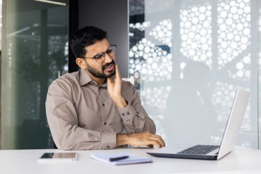 Çağdaş ofis ortamında dizüstü bilgisayar kullanırken diş ağrısı çeken bir adam. Yüzünde acı ve rahatsızlık var..