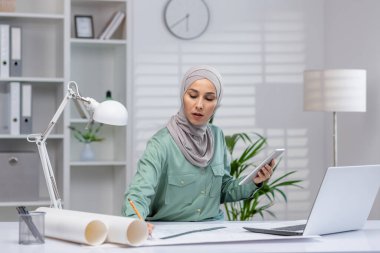 Türbanlı kadın mimar, çağdaş ofis ortamında tablet ve laptop kullanarak proje üzerinde çalışıyor.