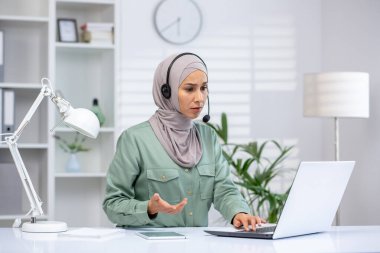 Kulaklık takan ve tesettürlü bir iş kadını modern bir ofiste sanal bir toplantı sırasında dizüstü bilgisayarla çalışıyor. Profesyonel, odaklanmış ve çevrimiçi bir tartışmayla meşgul..