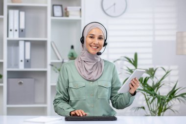 Neşeli müşteri hizmetleri temsilcisi kulaklık takıyor, elinde tablet tutuyor, modern bir ofiste çalışıyor. İş, iletişim ve destek temaları için ideal.