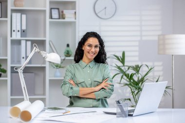 Modern ofis ortamında dizüstü bilgisayarı, belgeleri ve tasarım araçlarıyla masa başında duran kendinden emin bir kadın mimar..