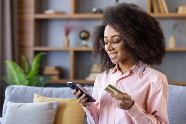 Gülümseyen bir kadın internet üzerinden kredi kartı ve akıllı telefonuyla ev ortamında rahat bir şekilde alışveriş yapar..