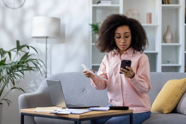 Genç bir kadın evdeki mali durumunu idare etmek için akıllı telefon ve laptop kullanıyor. Uzaktan çalışma ve mali yönetim kavramı.