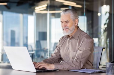 Modern bir ofiste dizüstü bilgisayarda çalışan kıdemli bir adam odaklanmayı ve üretkenliği gösteriyor. Cam duvarları ve doğal ışığı olan profesyonel çalışma ortamı.