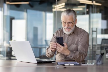 Üst düzey işadamı akıllı telefon ve dizüstü bilgisayar kullanarak ofiste oturuyor, çalışıyor ve iletişim kuruyor..