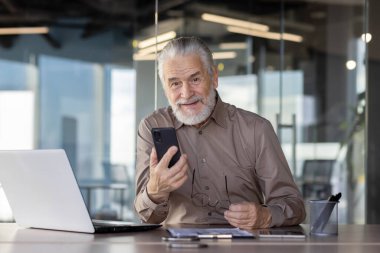 Modern ofiste bilgisayar ve cep telefonuyla çalışan kıdemli işadamı kameraya bakıyor ve gülümsüyor.