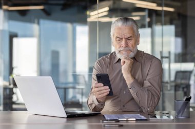 Modern bir ofiste çalışan yaşlı bir işadamı, önünde bir dizüstü bilgisayarla verileri analiz etmek için akıllı telefon kullanıyor.