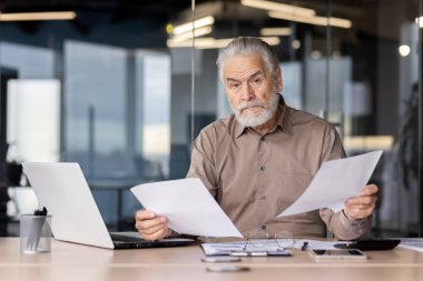 Gri sakallı kıdemli işadamı modern ofis ortamındaki belgeleri inceliyor, odaklanmış ve ciddi. Bilgisayarlı ve kağıtlı bir masada çalışmak..