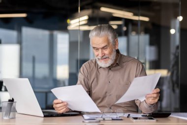 Modern bir ofiste kıdemli bir işadamı masasında dizüstü bilgisayarı ve okuma gözlükleriyle evrak işleriyle uğraşıyor.