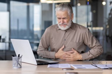 Modern ofis ortamında dizüstü bilgisayarda çalışırken mide ağrısı çeken yaşlı bir adam, sağlık ve rahatsızlığı vurguladı.