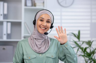 Türbanlı ve kulaklıklı genç Müslüman kadının yakın plan portresi modern parlak ofiste oturuyor, kameraya el sallıyor ve selamlıyor..