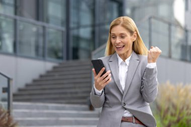 Ofis merkezinin yanında oturan mutlu genç iş kadını, telefon ekranına gülümseyerek bakıyor ve aldığı bilgiyle mutlu bir şekilde, zafer jestini eliyle göstererek kutluyor..