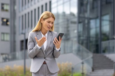 Gri takım elbiseli, üzgün genç bir iş kadını ofis binasının önünde duruyor ve cep telefonu ekranında hayal kırıklığına uğramış görünüyor..