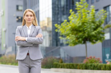 Bir ofis binasının önünde duran, kollarını göğsünün üzerinden geçirip kameraya bakan, kendine güvenen ve ciddi bir iş kadını portresi..