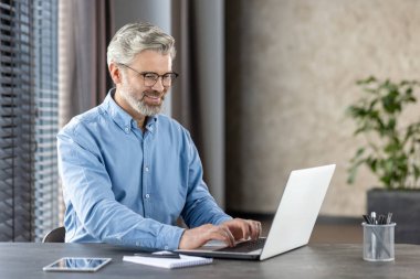 Gri saçlı ve sakallı olgun bir adam, ev ofis ortamında dizüstü bilgisayarla çalışıyor, kendine güvenini ve verimliliğini gösteriyor..