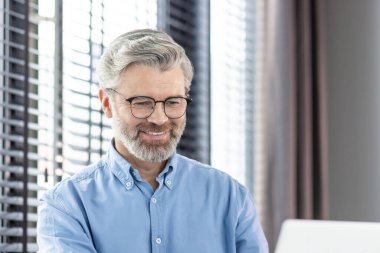 Gülümseyen, gözlüklü, mavi tişörtlü, arka planda beyaz perdelerle dizüstü bilgisayarda çalışan olgun iş adamı..