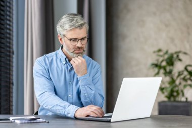 Masasında oturan gözlüklü olgun bir adam bilgisayarını kullanarak işine odaklanmış. Modern ofis ortamı.