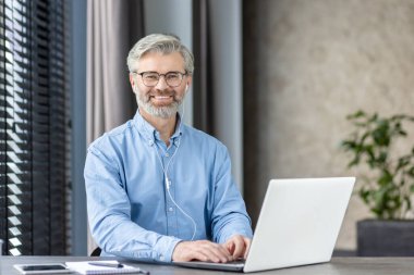 Modern ofis ortamında dizüstü bilgisayarda çalışan, güven ve profesyonellik saçan gözlüklü, gülümseyen olgun bir adam..