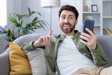 Kanepede oturan neşeli bir adam elinde akıllı bir telefonla video görüşmesi yapıyor ve neşeyle işaret ediyor. Arka plandaki iç mekan rahat bir atmosfer yaratıyor..
