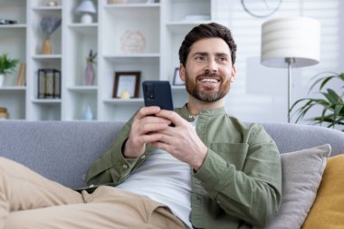 Akıllı telefon kullanarak gülümseyen, modern, şık bir oturma odasında koltukta dinlenen bir adam. Mutlu ve rahat görünüyor..