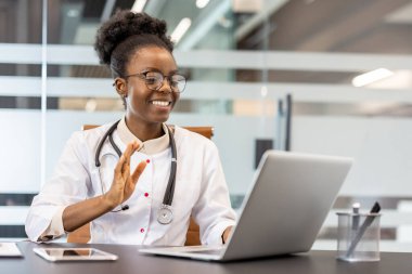 Steteskop takan gülümseyen bir doktor, modern bir ofiste dizüstü bilgisayarında bir hastayla sanal konsültasyon yapıyor..