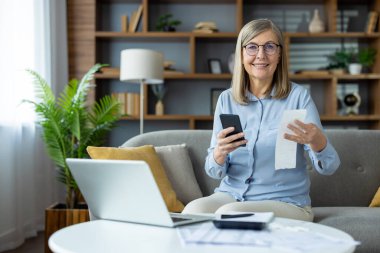 Gülümseyen ve elinde makbuz tutarken akıllı bir telefonla evde kişisel finans işlerini yöneten yaşlı bir kadın. Rahat ve rahat bir oturma odasında rahat bir atmosfer..
