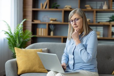 Evde dizüstü bilgisayar kullanan yaşlı kadın, derin düşüncelere dalmış. Uzaktan kumandalı profesyonel çalışma ve sorunları çözme.