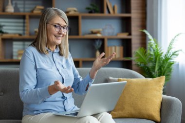Üst düzey bir kadın video görüşmesi sırasında konuşuyor, el kol hareketi yapıyor, dizüstü bilgisayar kullanıyor, rahat bir oturma odasında kanepede oturuyor.