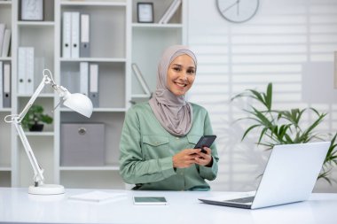 Başörtüsü takan gülümseyen profesyonel bir kadın, modern ofis ortamında bilgisayar, akıllı telefon ve masa lambasıyla çalışıyor..