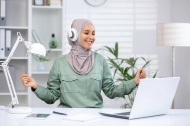 Kafiyeli mutlu bir kadın, kulaklıkla müzik dinlerken, modern ve parlak bir ofiste dizüstü bilgisayarla çalışıyor. Çoklu görev, keyif ve üretkenlik kavramı.